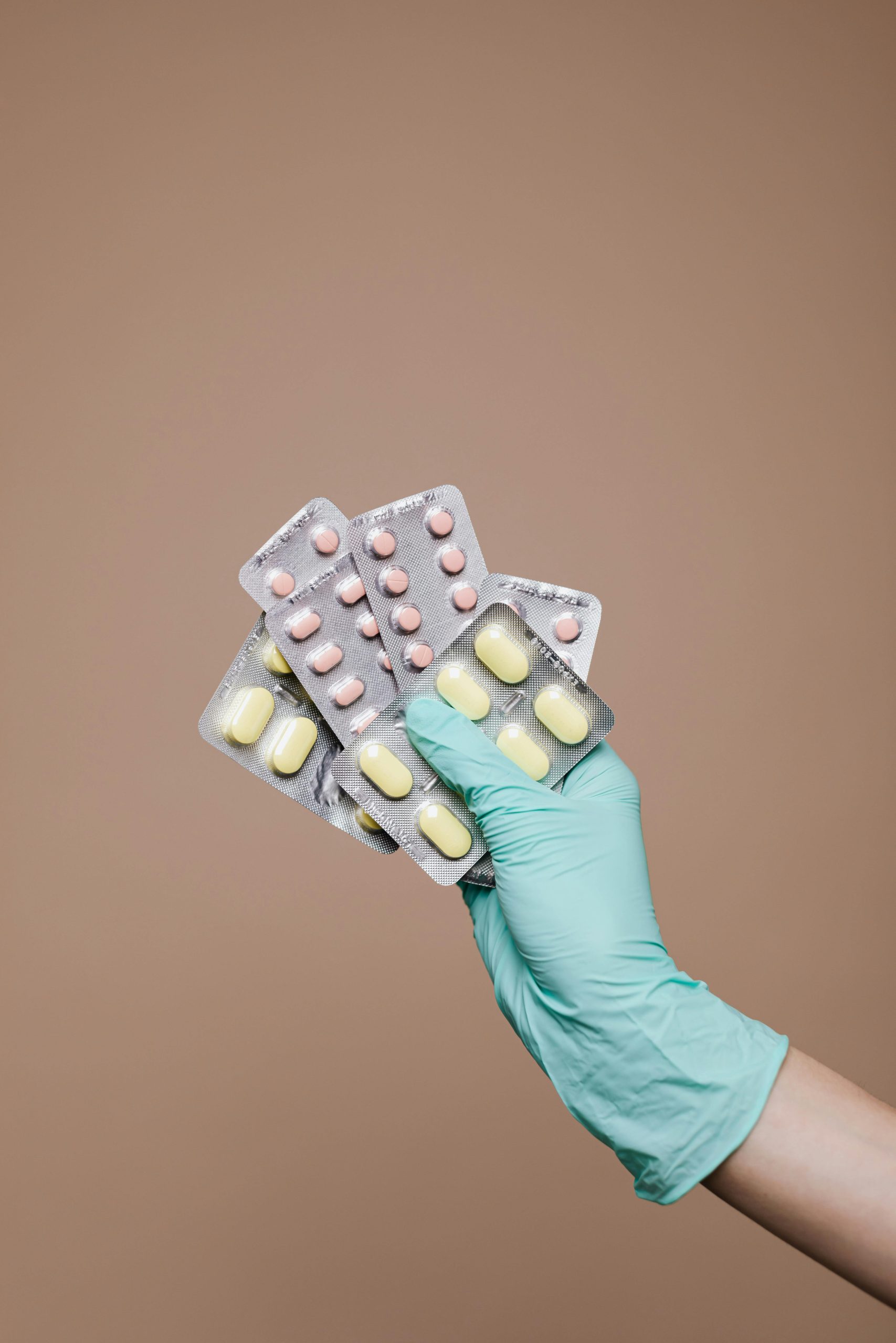 Close-up of a gloved hand holding various blister packs of pills against a neutral background.